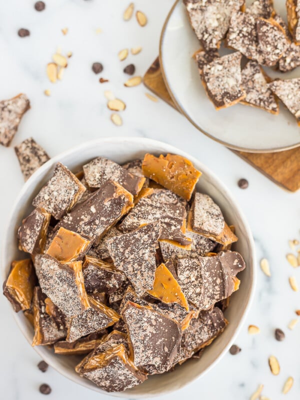 a bowl of homemade almond roca on a white board with almonds and chocolate chips