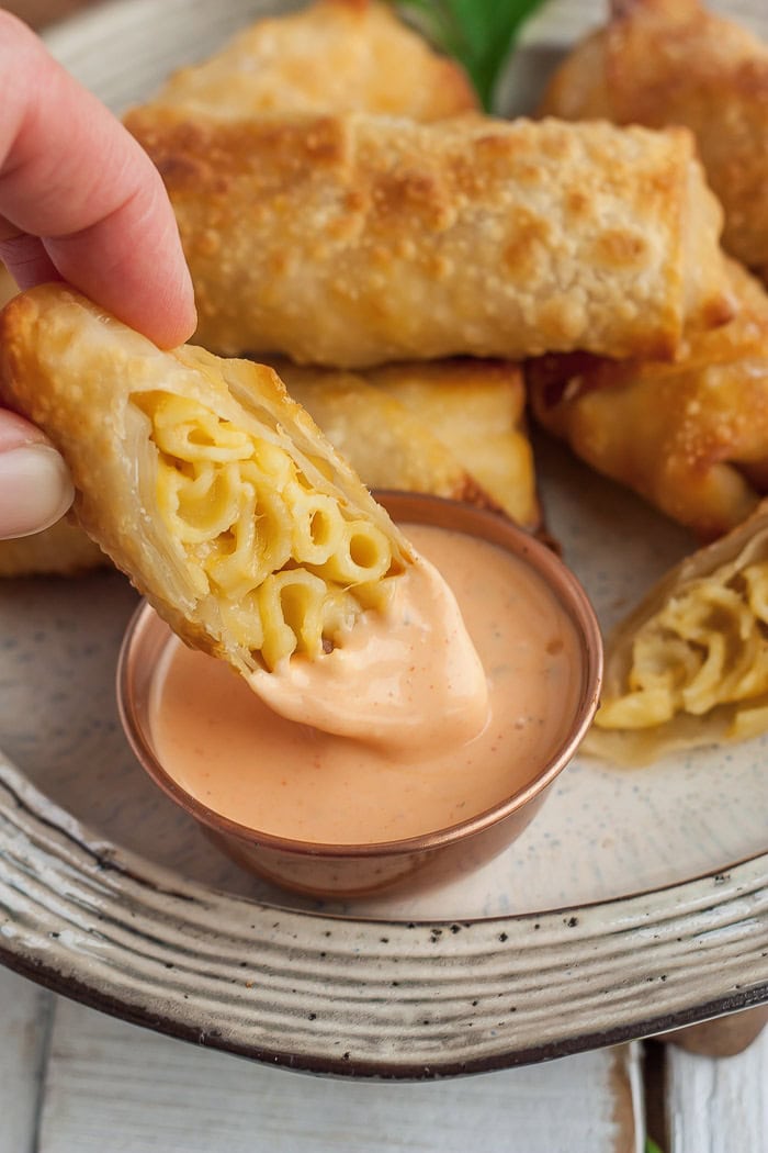 a hand dipping a mac and cheese egg roll into a bowl of buffalo ranch.