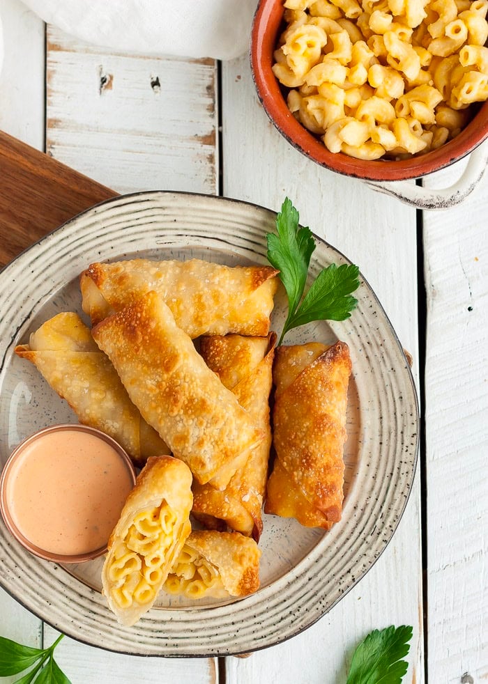 mac and cheese egg rolls on a beige plate with a small bowl of dipping sauce.