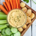 a glass bowl of cheese dip with veggies and pretzel bites on a wooden board
