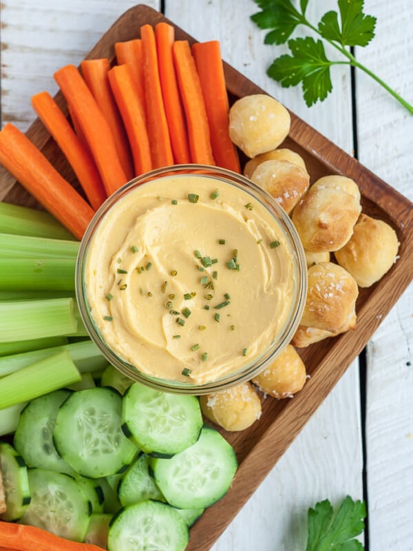 a glass bowl of cheese dip with veggies and pretzel bites on a wooden board