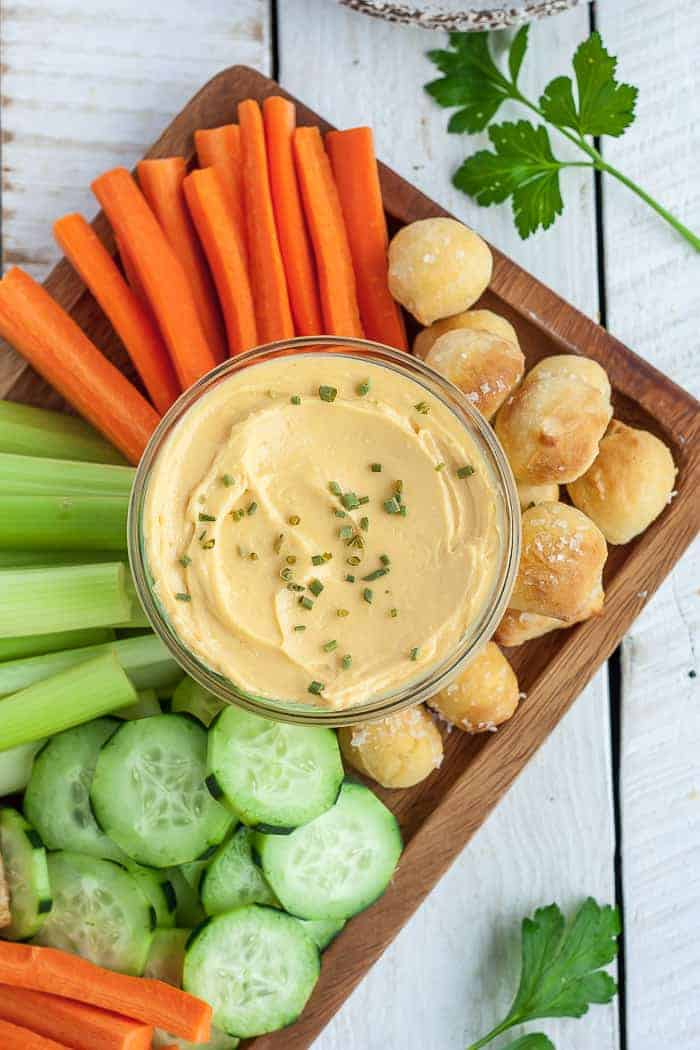 a glass bowl of cheese dip with veggies and pretzel bites on a wooden board