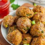 A white plate with Asian pork meatballs topped with diced onions with a small dish of chili sauce