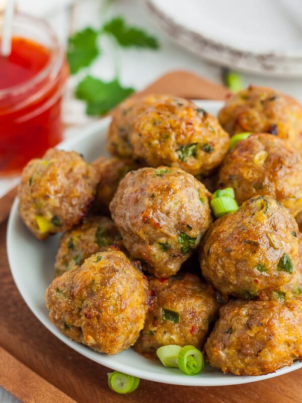 A white plate with Asian pork meatballs topped with diced onions with a small dish of chili sauce