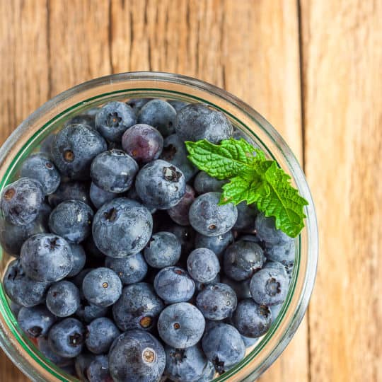 A glass bowl full of blueberries and a sprig of mint on a wooden board