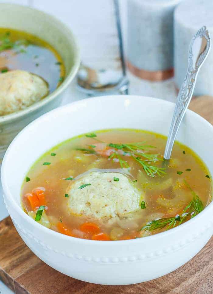 A white bowl of matzo ball soup with a spoon and fresh dill