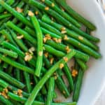 A close up look at a white bowl of green beans topped with garlic