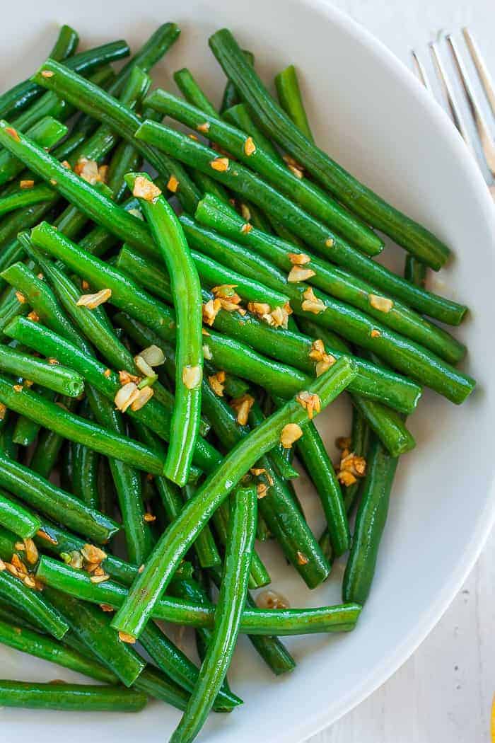 A close up look at a white bowl of green beans topped with garlic