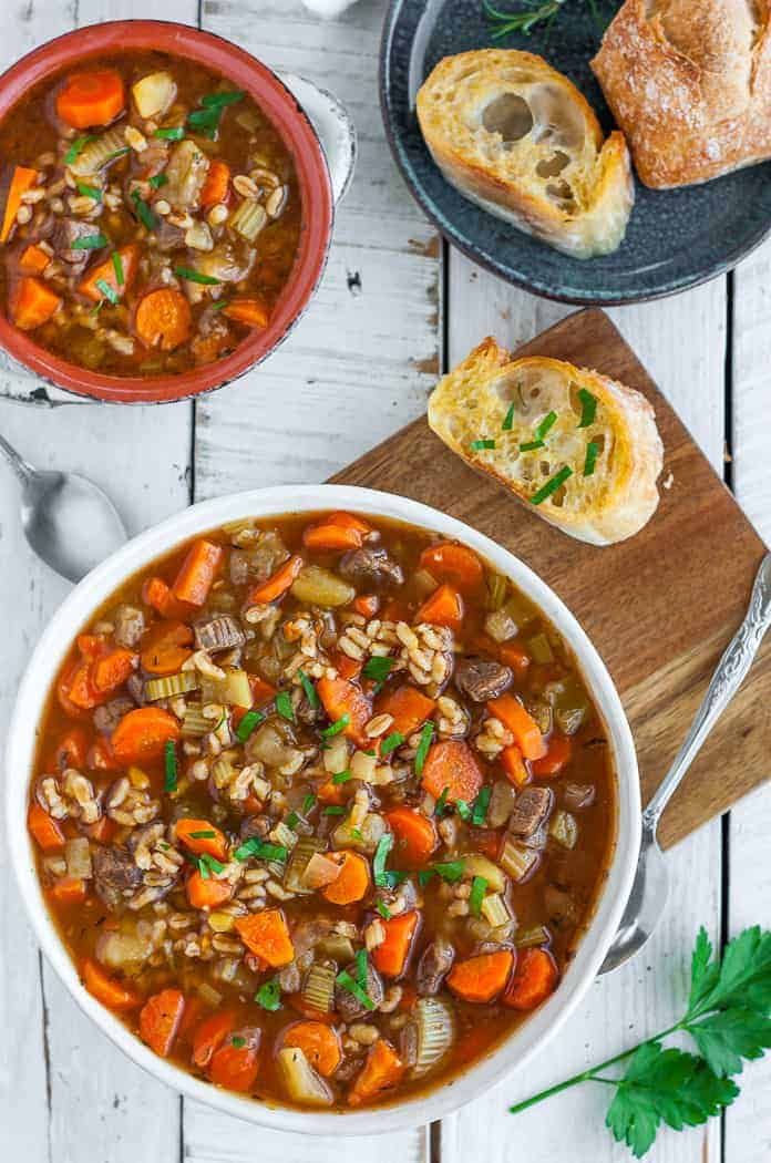 two bowls of Instant Pot Beef Barley soup on a board with sliced garlic bread.