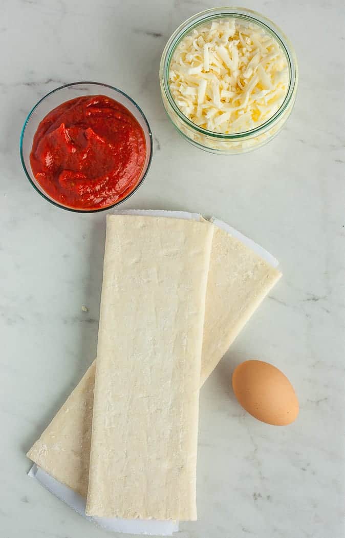 A sheet of frozen pastry, a dish of sauce and cheese, and an egg on a white piece of marble
