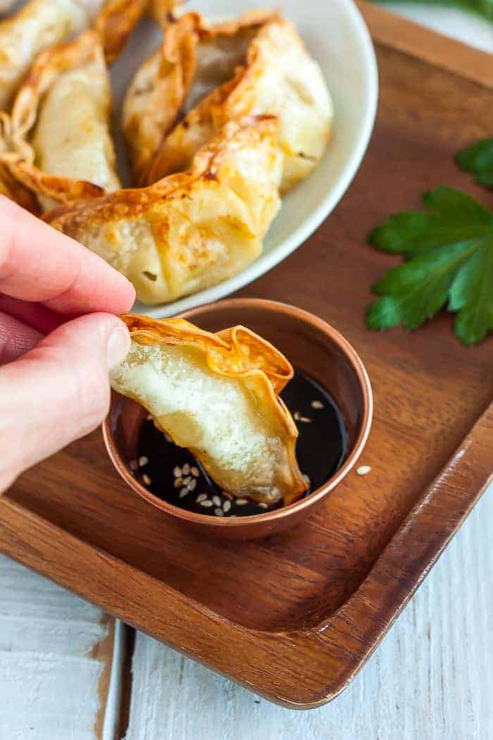 A hand dipping a crispy potsticker into a bronze bowl with sauce