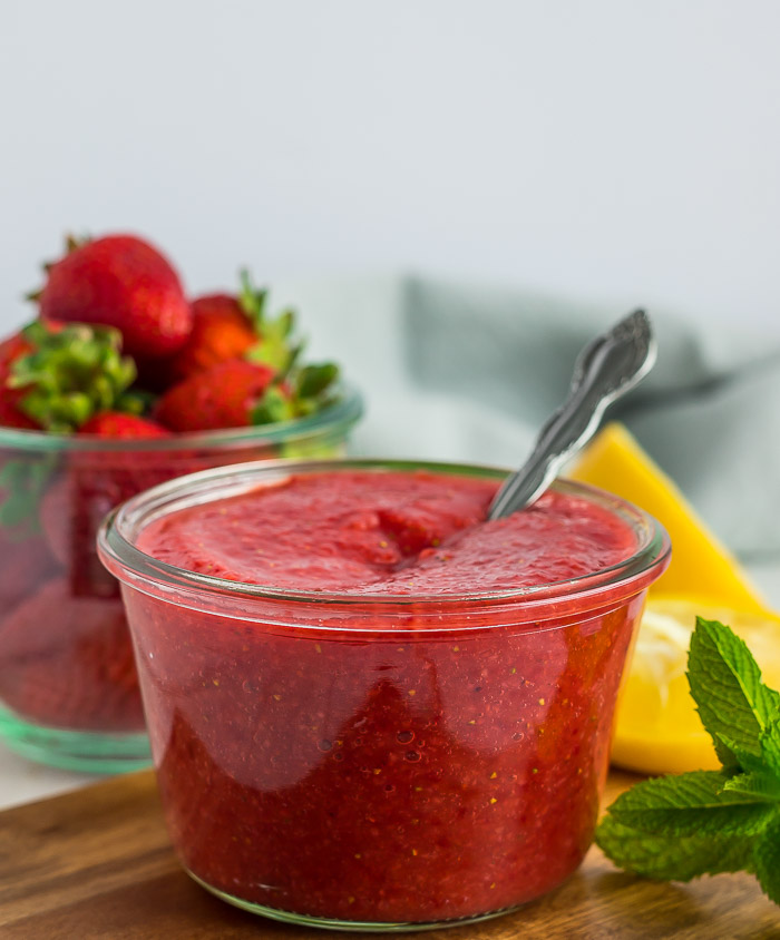 a glass jar of strawberry sauce with lemons and fresh strawberries on a wooden cutting board