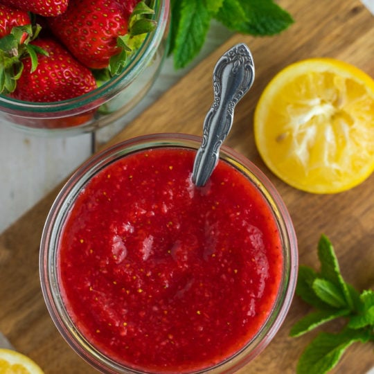 a glass jar full of strawberry sauce with a spoon, lemon, and mint in a wooden board