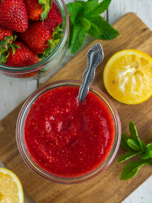 a glass jar full of strawberry sauce with a spoon, lemon, and mint in a wooden board