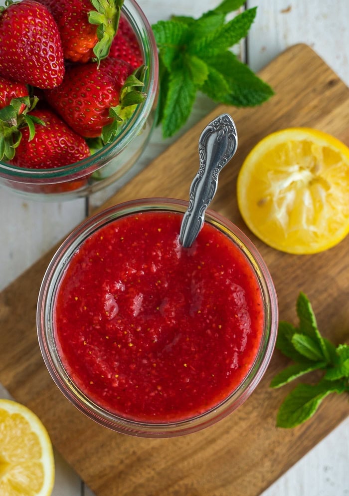 a glass jar full of strawberry sauce with a spoon, lemon, and mint in a wooden board