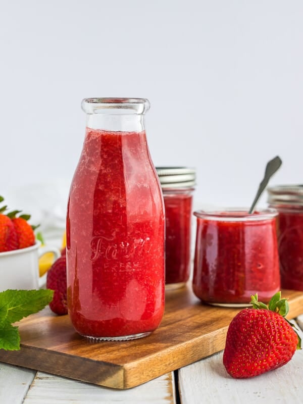 Mulitple glass jars of strawberry syrup on a wooden board with fresh strawberries and mint