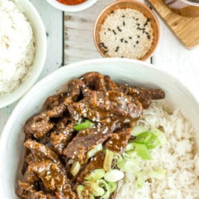 a white bowl of rice and sliced Instant Pot Beef Teriyaki topped with sesame seeds and green onions.