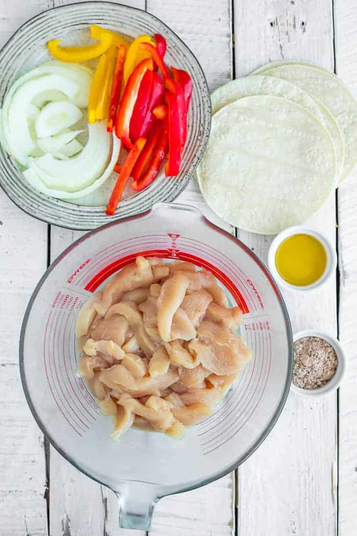 a bowl of sliced chicken, a plate of onions, and peppers, and bowls of seasoning on a white board