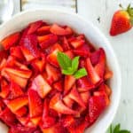 A bowl of sugared straweberries topped with mint on a white board