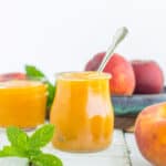 a glass jar of peach syrup with a spoon in it on a white wooden board