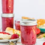 Multiple jars of strawberry rhubarb jam on a white board and a biscuit