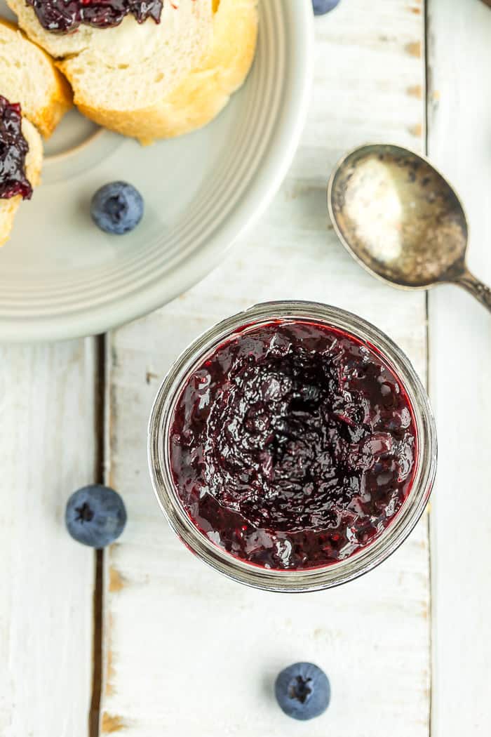 a close up photo of a glass jar of blueberry freezer jam on a white board with a spoon