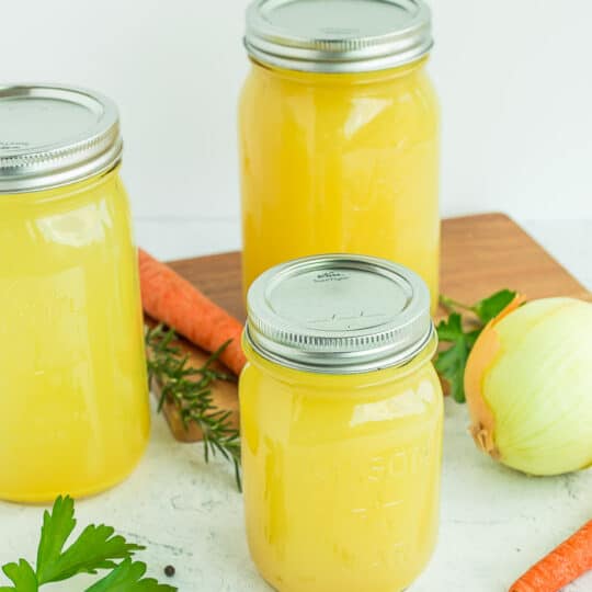 mason jars of chicken stock on a white surface with veggies and herbs