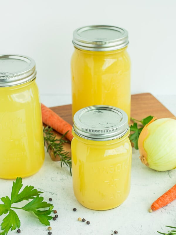 mason jars of chicken stock on a white surface with veggies and herbs