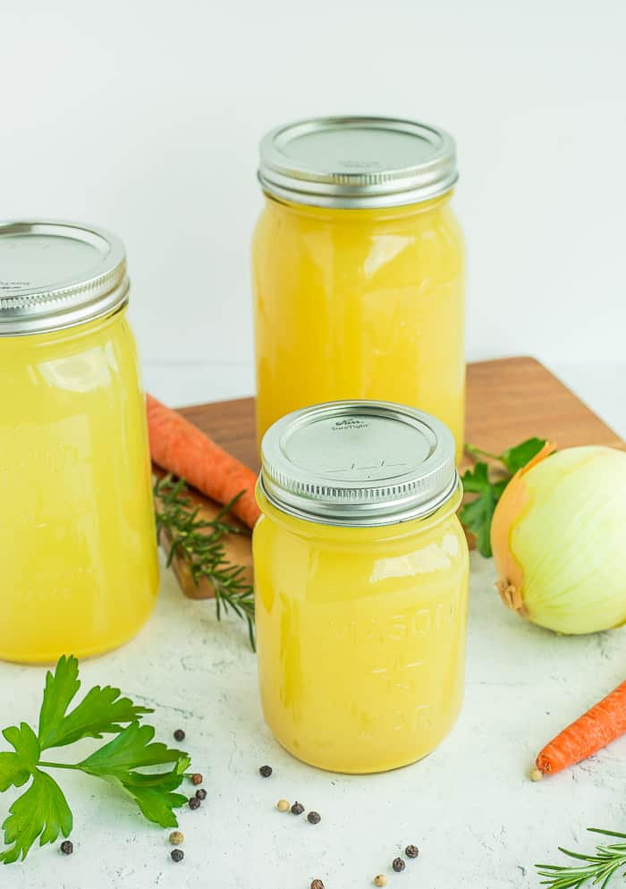mason jars of chicken stock on a white surface with veggies and herbs