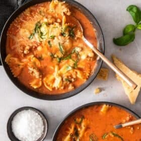 two bowls of lasagna soup on a white surface with basil and cheese
