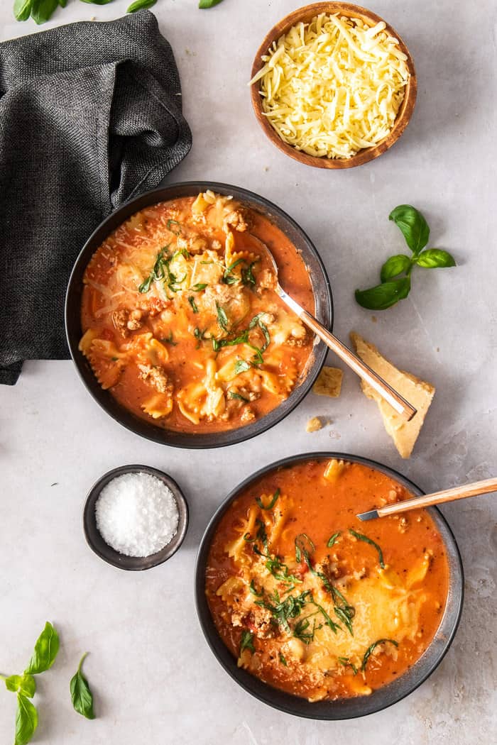two bowls of lasagna soup on a white surface with basil and cheese