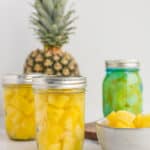3 pint jars of canned pineapple on a white board with a full pineapple and a bowl of pineapple