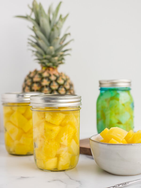 3 pint jars of canned pineapple on a white board with a full pineapple and a bowl of pineapple