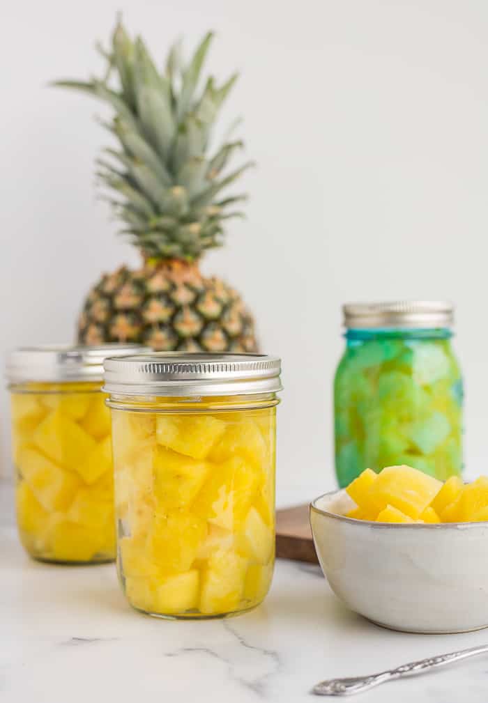 3 pint jars of canned pineapple on a white board with a full pineapple and a bowl of pineapple