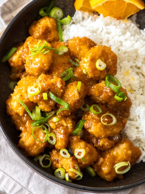 a bowl of air fryer orange chicken topped with green onions in a bowl with white rice and orange slices