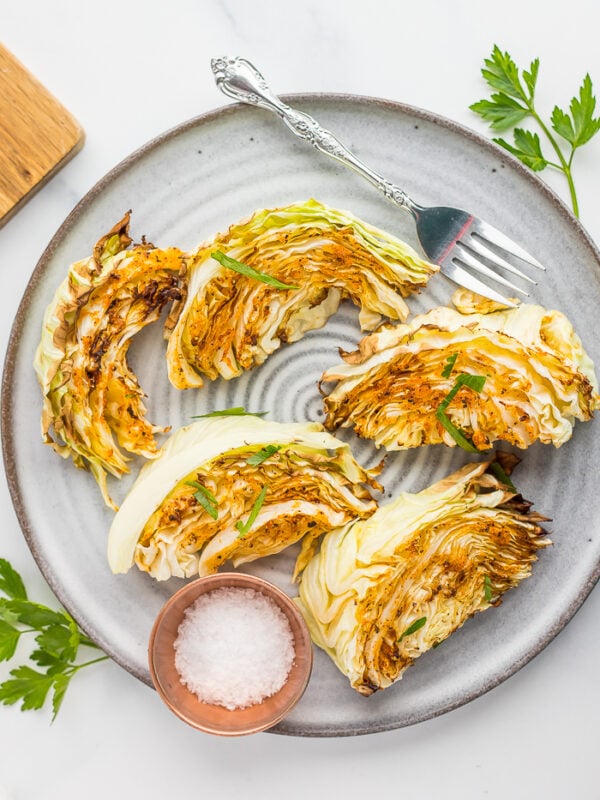 5 roasted cabbage wedges on a grey plate with a fork and a small bowl of kosher salt.