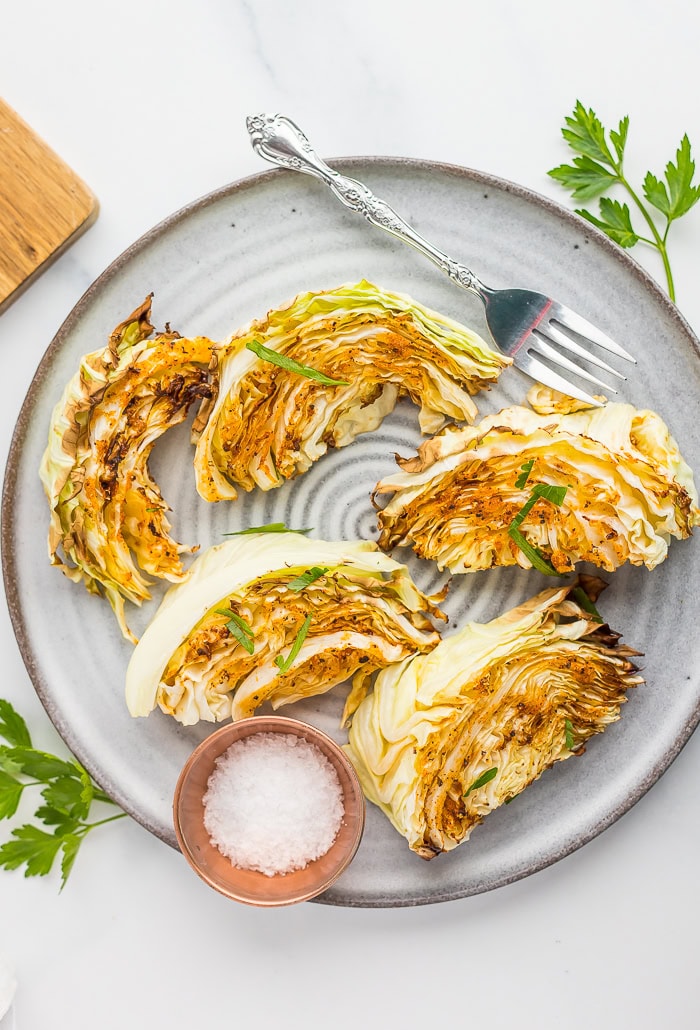 5 roasted cabbage wedges on a grey plate with a fork and a small bowl of kosher salt.