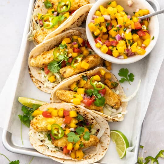 crispy air fryer fish tacos in tortillas on a tray with a bowl of mango salsa