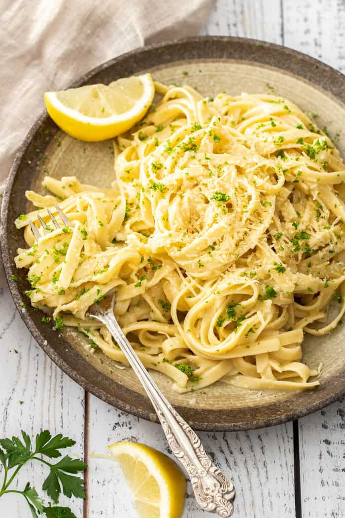 A plate of lemon garlic pasta topped with Parmesan cheese, parsley, and a slice of lemon.