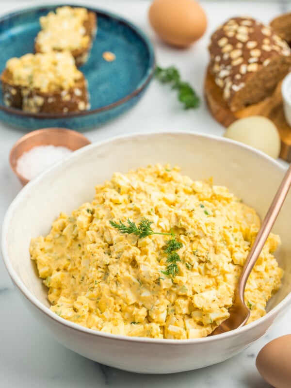 mayo free egg salad in a white bowl with brown bread and eggs on a white board