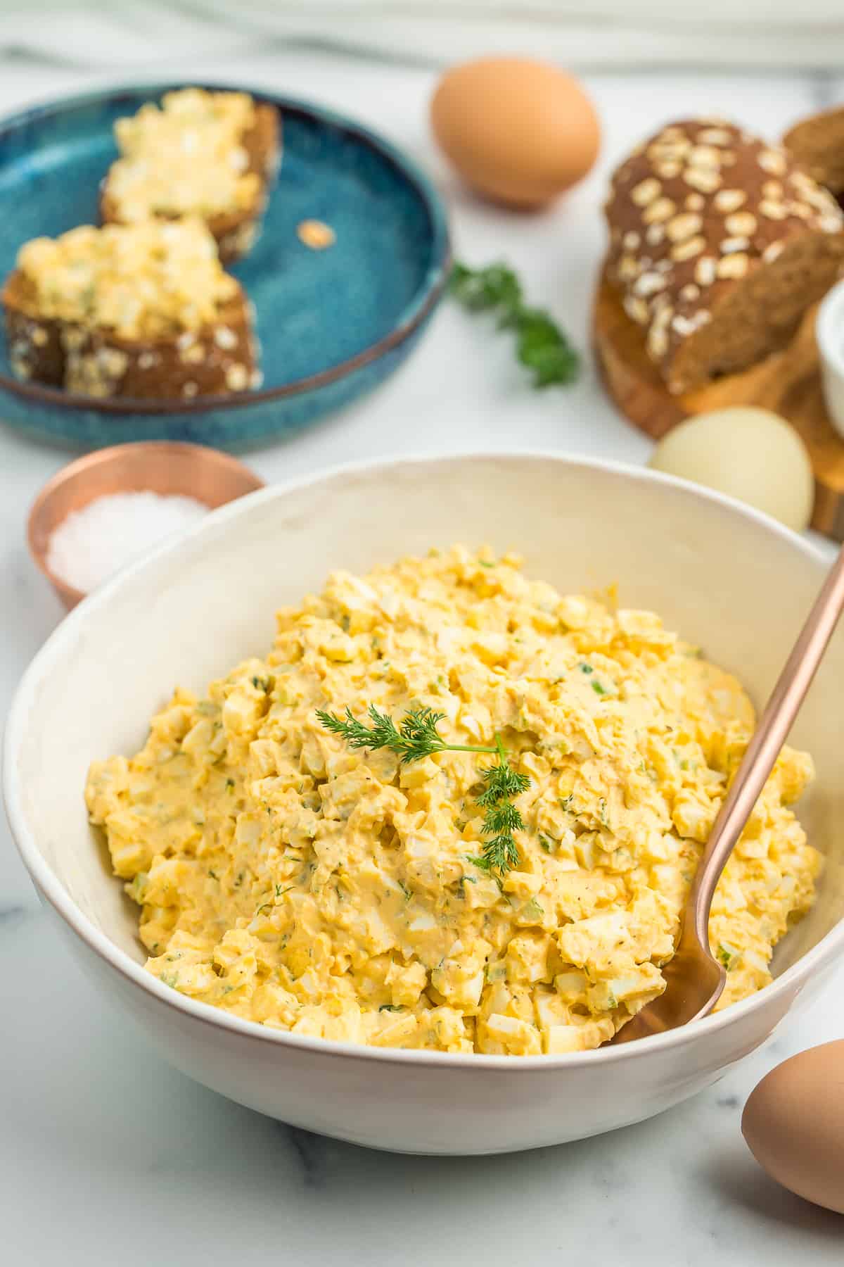 mayo free egg salad in a white bowl with brown bread and eggs on a white board