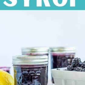 glass jar of blackberry syrup with fresh blackberries and a lemon on a white board.