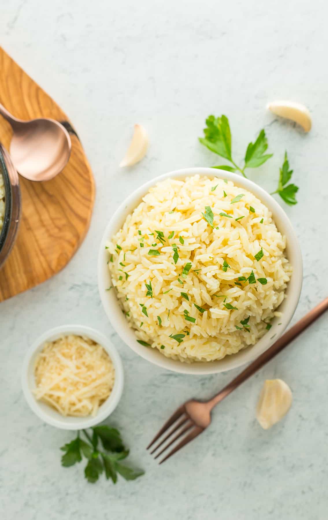 bowls of garlic parmesan rice on a white board with parsley and garlic bulbs.