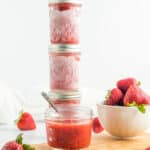 stacked jars of strawberry freezer jam with fresh strawberries on a white board.