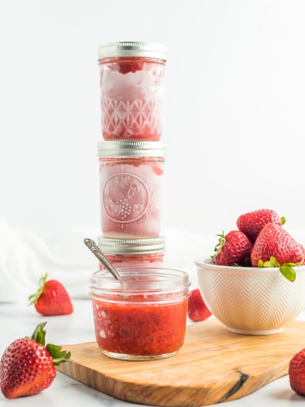 stacked jars of strawberry freezer jam with fresh strawberries on a white board.