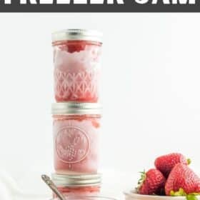 stacked jars of strawberry freezer jam with fresh strawberries on a white board.