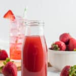 a glass bottle with strawberry simple syrup with fresh strawberries and a glass behind it.