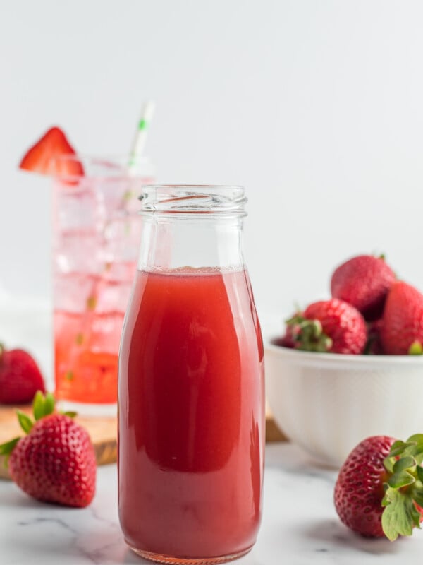 a glass bottle with strawberry simple syrup with fresh strawberries and a glass behind it.