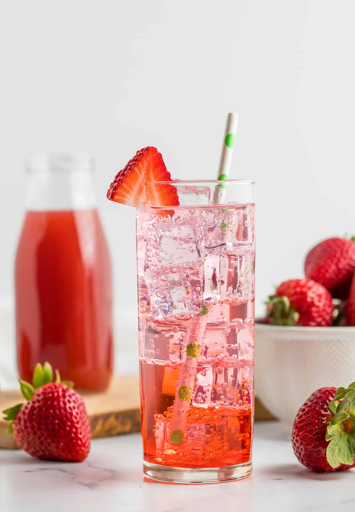 a glass of sparkling water and strawberry syrup for drinks with a green straw and sliced strawberries