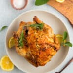 a roasted air fryer cornish hen on a white plate with sage, lemon, and a bowl of salt.